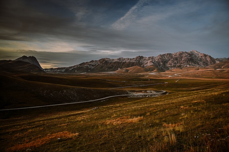 A hiker walks along a rocky trail through high-altitude grasslands with the Gran Sasso mountain peaks in the background.